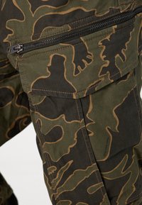 Stof met camouflagepatroon in olijfgroene en bruine tinten; heeft een zichtbare ritszak en een gestructureerd oppervlak.