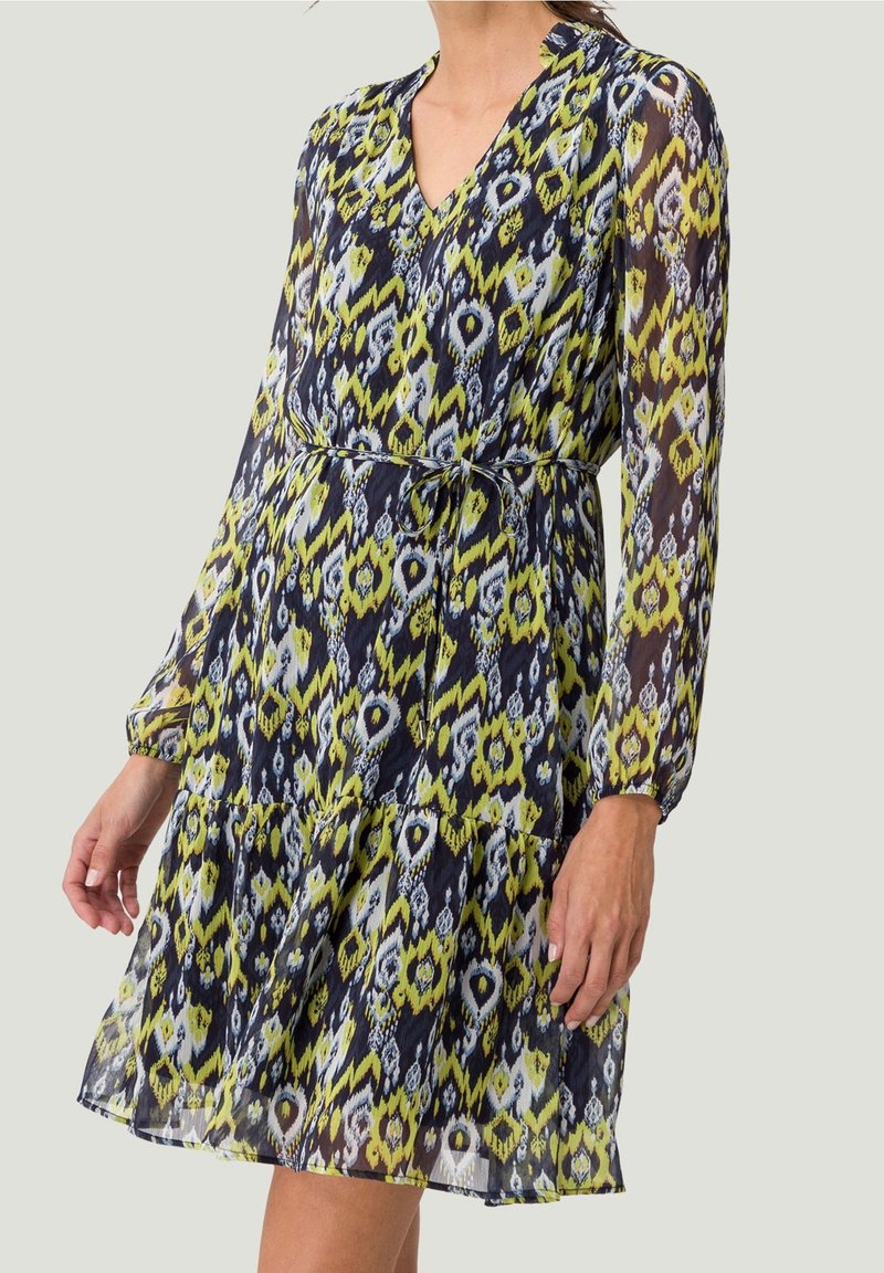 Femme portant une robe à manches longues, de longueur genou, avec un motif géométrique abstrait bleu, jaune et blanc, et une fine ceinture à la taille.