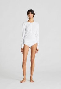 Bílý top s dlouhým rukávem a henley výstřihem, opatřený knoflíkovou légou se třemi knoflíky, v kombinaci s bílými krajkovými kalhotkami. Měkká, hladká tkanina, těsné provedení.