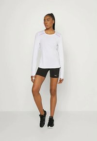 Weißes langärmliges Sportoberteil mit rosa Akzenten, kombiniert mit schwarzen Shorts. Das Model trägt schwarze Sportschuhe und steht in einer neutralen Pose.