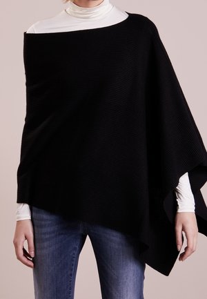 Persoon draagt een zwarte asymmetrische poncho over een witte coltrui en blauwe spijkerbroek, staand tegen een effen achtergrond.