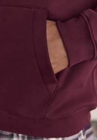 Burgunderröd huvtröja med känguruficka, ribbade ärmslut och nederkant. Tyget verkar mjukt med en slät yta och en avslappnad passform.