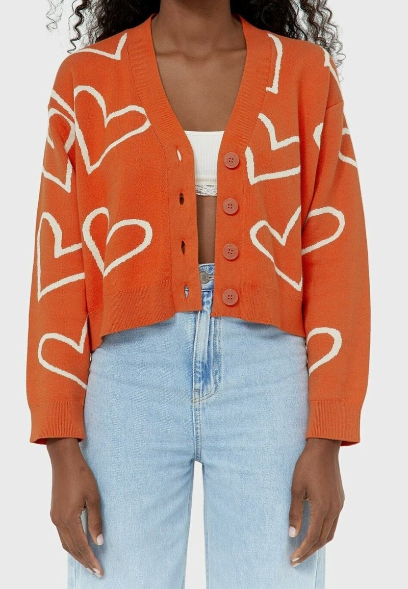 Cardigan court orange avec motifs de cœurs blancs, quatre boutons, porté sur un haut court blanc et un jean taille haute bleu clair.