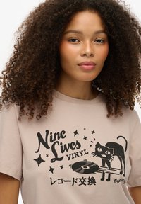 Lysebeige bomulds t-shirt med sort tryk af en kat, der leger med en vinylplade, med dekorative stjerner og textelementer.