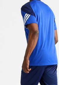 Sininen urheilu paita, jossa tummansiniset yksityiskohdat ja kolme valkoista raitaa olkapäällä. Sileä kangas, lyhyet hihat ja pyöreä kaula-aukko.