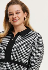 Schwarz-weißes Hahnentritt-Musterkleid mit schwarzem Kragen, ausgestattet mit einem zentralen Reißverschluss und strukturiertem Stoff.
