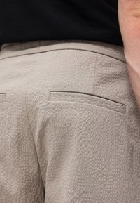 Κοντινό πλάνο μπεζ παντελονιού με υφή και ραμμένες πίσω τσέπες, φορεμένο από άτομο με μαύρο τοπ, δείχνοντας μέρος του μπράτσου του.