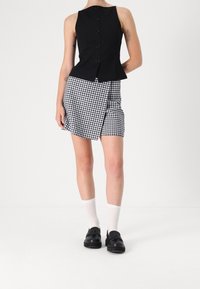Top senza maniche nero con bottoni sulla schiena, abbinato a una gonna avvolgente a scacchi bianco e nero. Il modello indossa scarpe nere e calzini bianchi.