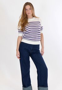 Pikkade juustega naine, kellel on seljas valge lühikeste varrukatega kootud topp lilla triipudega ja tumedad laiad püksid, seisab ühevärvilise tausta ees.