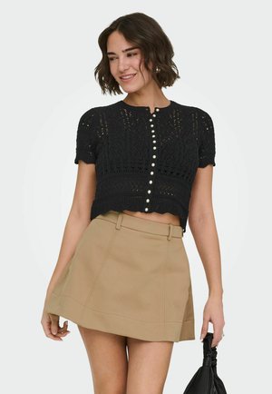 Kvinne som smiler, iført en svart kortermet bluse med knapper i blonder og beige miniskjørt, holder en svart sammenrullet håndveske.