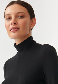Črna rebrasta turtleneck majica z oprijetim dizajnom in gladko teksturo. Ponuja visok ovratnik in je poudarjena z zlatimi visečimi uhani.