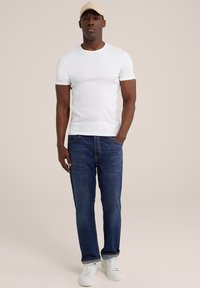 Weißes T-Shirt mit kurzen Ärmeln, kombiniert mit dunkelblauen Jeans und weißen Sneakers. Die Person steht ruhig da, die Hände in den Taschen, und trägt eine beige Kappe.