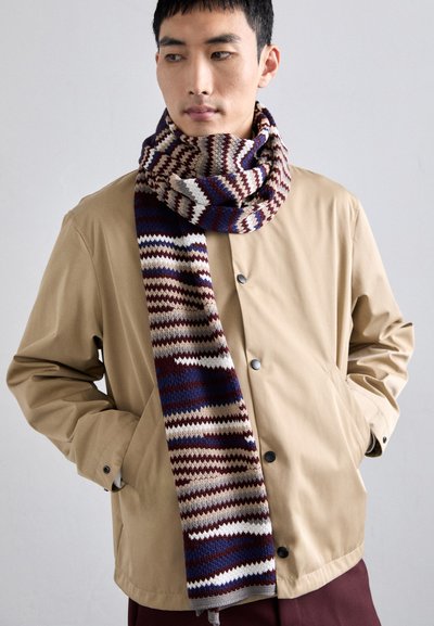Sciarpa lavorata a maglia con un motivo a zigzag nei colori marrone, crema e blu, avvolta attorno al collo, abbinata a una giacca beige e pantaloni bordeaux.