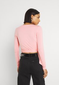 Top de mangas longas cor-de-rosa com um design ajustado, apresentando um detalhe de costas torcido, combinado com jeans pretos de cintura alta e um cinto brilhante.