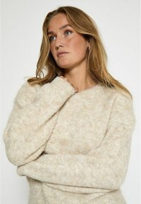Beżowy dzianinowy sweter o miękkim, teksturowanym wykończeniu, z okrągłym dekoltem i długimi, luźnymi rękawami. Materiał wydaje się przytulny i ciepły.