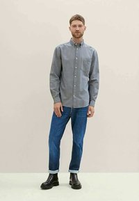 Camisa cinza de botão com um bolso no peito, combinada com jeans azuis e botas pretas. A camisa tem colarinho e mangas longas.