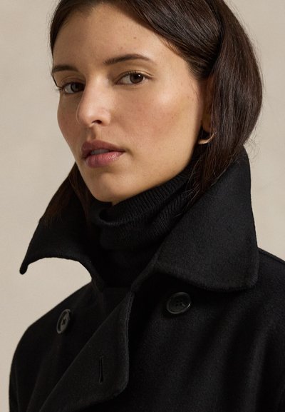 Černý dvouřadý kabát s vysokým límcem, vyrobený z texturované tkaniny. Obsahuje velké černé knoflíky a pod ním je vrstvený rolák.