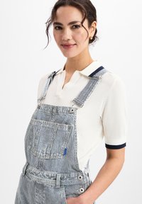 Vrouw in een witte poloshirt met korte mouwen en blauwe en witte gestreepte denim overall, lachend met haar hand in de zak tegen een witte achtergrond.