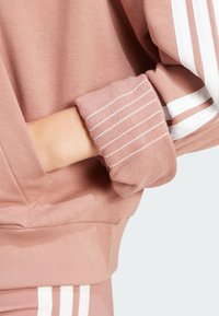 Roza kapuca s bele črtaste podrobnosti na manšetah, mehka tekstura in sproščen kroj z vidno žepom na strani.