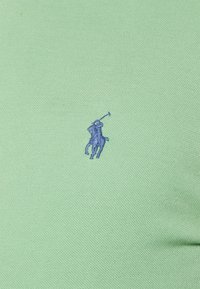 Niebieskie haftowane logo polo na jasnoszarym teksturowanym tle.