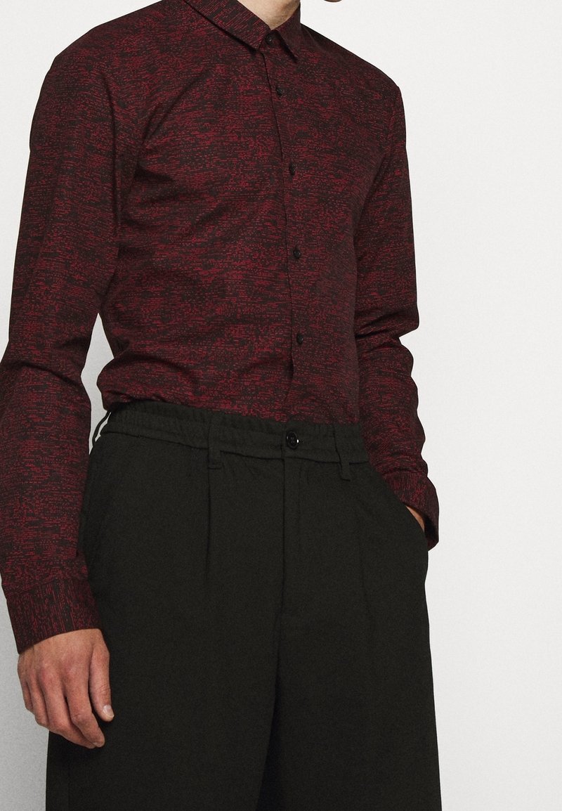 Camicia a maniche lunghe rossa con texture, colletto button-down e bottoni neri, abbinata a pantaloni neri con vestibilità comoda e tasche visibili.
