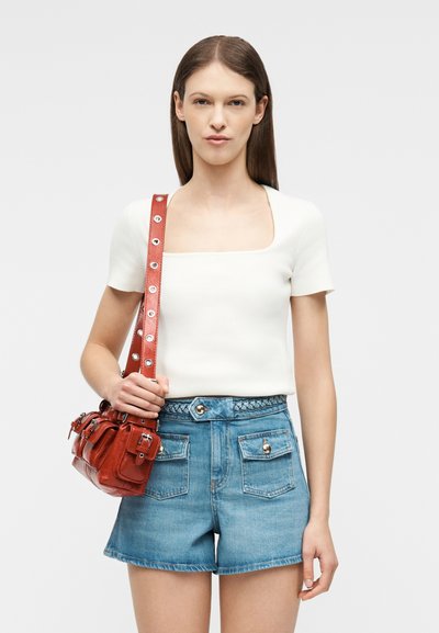 Γυναίκα που φοράει λευκό τοπ με κοντά μανίκια και τζιν σορτς ψηλόμεσο με πλεκτή ζώνη, κρατώντας κόκκινη δερμάτινη τσάντα ώμου με μεταλλικά κουμπώματα.