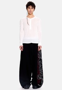 Ilgomis rankovėmis balti henley marškiniai su sagų dygsniu, derinami su laisvais juodais kelnėmis, šonuose puoštomis raudonais ir baltais sūkurėlių raštais.