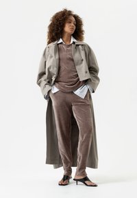 Grijze trenchcoat over een bruin fluwelen top en broek, met daaronder een blauw-wit gestreept shirt; zwarte pumps maken de look af.