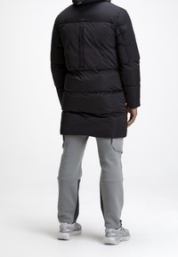 Manteau matelassé noir avec capuche, doté d'un détail zippé dans le dos, accompagné d'un pantalon de sport gris et de baskets grises.