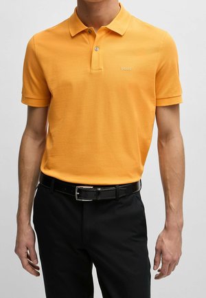 Mies, joka käyttää kirkkaan oranssia lyhythihaista poolopaitaa, jossa on "BOSS"-logo, ja mustia housuja, joissa on musta nahkavyö.