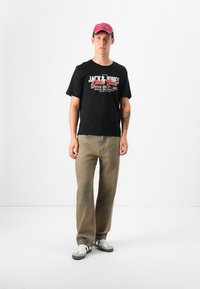 En ung mann står mot en hvit bakgrunn, iført en svart t-skjorte, khaki-bukser og en rosa caps, med hvite og grå sneakers.