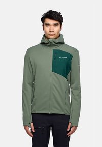 Giacca softshell verde con tasca sul petto con cerniera, cappuccio e pannello verde scuro a contrasto sulla parte superiore sinistra.