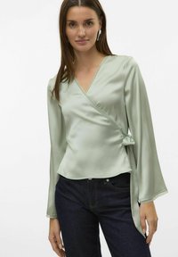Ljusgrön omslagstopp i slät tyg med ett knytband, scalloped spetsdetaljer längs halsringningen och ärmarna, tillsammans med mörka jeans.