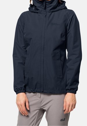 Femme portant une veste imperméable noire avec capuche et un pantalon gris clair, debout devant un fond blanc uni.