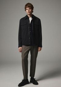 Sort jakke med brystlommer, parret med en marineblå skjorte med knapper, grå bukser og sorte snøresko på en lys baggrund.