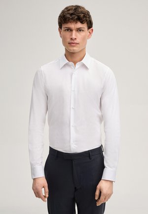 Młody mężczyzna o krótkich brązowych włosach, ubrany w dopasowaną białą koszulę na guziki z długim rękawem oraz ciemne spodnie, na jednolitym tle.