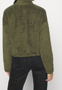 Chaqueta de forro polar texturizada de color verde oliva con cuello alto y diseño corto, llevada sobre unos vaqueros negros. Sin cierres ni patrones visibles.