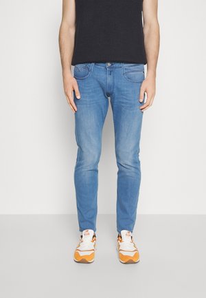 Homme portant un jean slim bleu clair, un top noir sans manches et des baskets blanches avec des accents orange, debout sur un fond blanc.