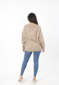 Beige oversized gebreide trui met geribbelde manchetten en zoom, gedragen bovenop strakke blauwe spijkerbroeken, gecombineerd met doorzichtige haksandalen.