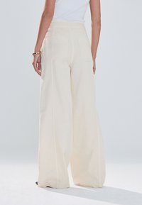 Pantalon crème à jambes larges en tissu doux. Il présente une taille haute, deux poches arrière et des détails de couture apparents.