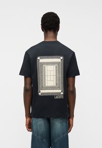 Άνδρας που φ οράει μαύρο μπλουζάκι Lacoste με λευκό ορθογώνιο γεωμετρικό σχέδιο στην πλάτη, συνδυασμένο με μπλε τζιν, στέκεται μπροστά σε απλό φόντο.