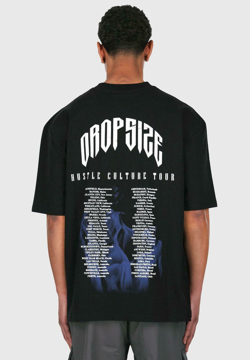 Sort oversized t-shirt med fed hvid tekst "DROPSIZE" og "HUSTLE CULTURE TOUR" samt en liste over steder trykt i hvid på bagsiden.