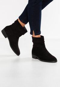 Bottes de cheville en daim noir au design flou, avec un bout rond et un petit talon carré, associées à un jean en denim foncé.