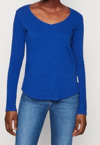 Nainen, jolla on päällä kirkkaan sininen pitkähihainen V-kauluspaita ja vaaleansiniset farkut, seisoo yksinkertaista valkoista taustaa vasten.