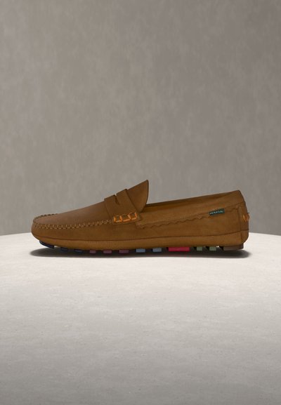 Mocassin en daim marron avec détails cousus et semelle en caoutchouc multicolore, présenté sur une surface claire contre un fond beige uni.