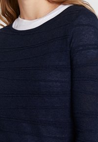 Granatowy sweter dzianinowy z teksturowanymi poziomymi paskami, okrągłym dekoltem i lekkim materiałem. Widoczny jest ecru detal wewnętrznego kołnierza.