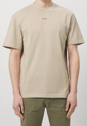 Beżowy T-shirt z krótkim rękawem z logo "HUGO" na piersi, noszony przez osobę w oliwkowozielonych spodniach, stojącą na tle jednolitego jasnego tła.