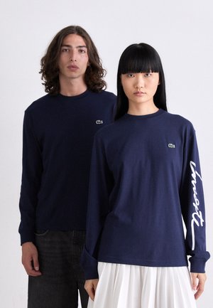 Tamsiai mėlynos ilgomis rankovėmis marškiniai, pagaminti iš medvilnės, su mažu žaliu logotipu krūtinėje ir baltu tekstu ant vienos rankovės. Unisex dizainas.