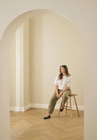 Moteris ilgais rudais plaukais, vilkinti baltą megztinį ir alyvuogių spalvos kelnes, sėdi ant medinės kėdės minimalistiškame kambaryje su arkiniais durimis.