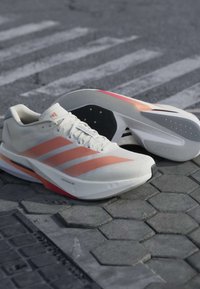 Ζευγάρι άσπρα και πορτοκαλί παπούτσια Adidas για τρέξιμο ακουμπισμένα σε έναν πλακόστρωτο δρόμο με διάβαση πεζών στο βάθος.